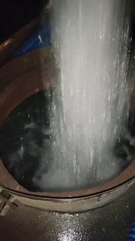 A high-angle shot shows a large volume of white foamy water being discharged from a blue ribbed hose into a circular metal vat. The water is cascading forcefully, creating turbulence and splashes within the vat, which contains dark liquid. The hose is positioned vertically above the vat and appears to be delivering a continuous flow. The vat has a thick rim and is partially submerged in what appears to be a concrete or paved surface. The lighting is dim, with the water stream being the primary source of illumination. The scene suggests an industrial or construction-related activity involving liquid transfer or disposal.