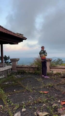 A person with a colorful jacket, dark pants, and purple shoes stands on an overgrown, partially collapsed, roof or terrace taking pictures with a smartphone of the distant landscape under a cloudy sky. Fragments of red tiles are scattered across the ground, along with weeds and other debris. There is a partially collapsed structure in the background with an intact roof. The landscape in the distance consists of rolling hills and a valley under an overcast sky.