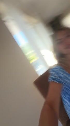 The video begins with a close-up, selfie-style shot of a woman with blonde hair, wearing a blue floral dress. She is looking at the camera and appears to be singing or talking, with her mouth open. The camera is angled downwards, showing her torso and lap. In the background, another person, a younger female with dark hair wearing a white t-shirt and pink shorts, is sitting at a table, also looking away from the camera and speaking. The table is set with food, including what appears to be shrimp, bread, and fruit, as well as drinks. The environment looks like an indoor dining area or kitchen, with cabinets and a doorway visible in the background. The lighting suggests it is daytime. The overall scene is casual and appears to be a candid moment.