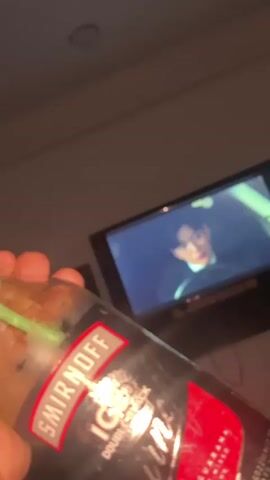 The video shows a person holding a Smirnoff Ice Double Black Guarana alcoholic beverage in their hand. A green straw is visible in the bottle. In the background, a television is on, displaying a scene from a show with a man in glasses lying on a bed and looking at his phone. Subtitles are visible on the screen. The room appears dimly lit.