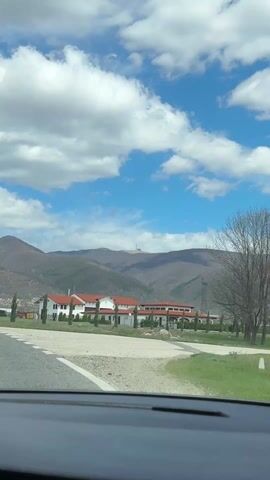 The video appears to be taken from inside a moving vehicle, possibly a car, driving on a paved road towards a building complex. The complex consists of multiple buildings with white walls and red roofs, situated in a flat area. The focus shifts to a mountain range in the background, on top of which stands a prominent structure. The structure is a large, round building with a tall spire extending upwards, seemingly isolated on the mountain peak. The sky is blue with scattered white clouds, and the weather seems clear. There are trees and power lines visible near the complex and in the foreground. The camera zooms in slightly on the structure on the mountain.