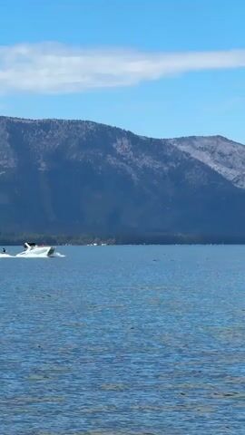 A white speedboat is moving left to right across a blue lake. There are people in the boat, and one person is water skiing behind it. The lake is calm with gentle ripples. In the background, there are dark green trees and large, gray mountains under a bright blue sky with a few white clouds.