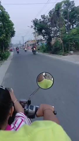 The video is shot from the perspective of someone riding on the back of a motorcycle, looking forward. The rider's hands are visible on the handlebars. In front of them, a child is seated on the motorcycle, wearing a striped shirt. A motorcycle mirror reflects the face of the person driving the motorcycle, who is wearing a bright green shirt. The motorcycle is on a paved road, with other motorcycles and people visible in the distance. Buildings line the side of the road, and there are power lines overhead. The sky is overcast, suggesting it might be dawn or dusk.