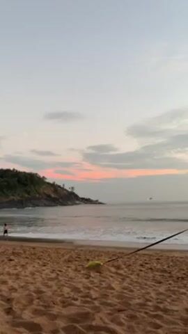 The video shows a beach scene at dusk or dawn. The sand is a reddish-brown color. Gentle waves roll onto the shore. A headland covered in trees juts out into the ocean on the left side of the frame. In the distance, the sky is a pale pink and grey, suggesting either sunrise or sunset. A person is faintly visible walking on the beach near the water's edge on the left side. A black leash is stretched from the foreground to the water. A green ball or toy sits on the sand in the lower left.