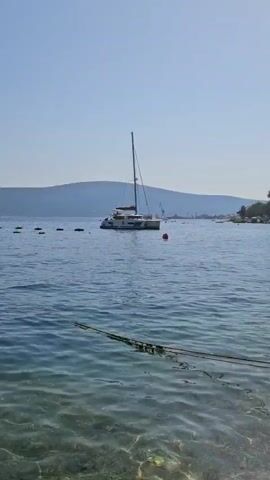 The video shows a catamaran anchored in calm, blue water. A red buoy is visible near the boat. In the background, a hilly coastline with some industrial structures and cranes can be seen. The sky is clear and blue. The foreground shows a dark rope extending into the water.