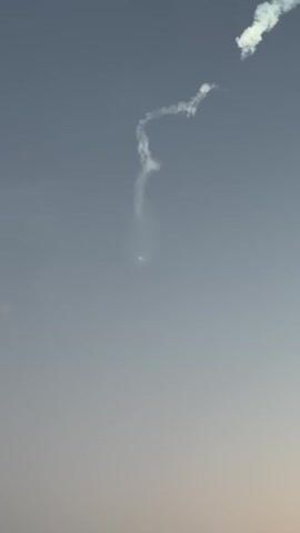 The video shows a clear, pale blue sky with a few wispy clouds. In the upper right corner, a distinct white smoke trail curves and then dissipates. Below this, a similar, but more elongated smoke trail descends from the upper portion of the frame. Near the center of the frame, a bright, small, luminous object is visible, emitting a subtle glow. This object appears to be the source of the descending smoke trail. As the video progresses, the luminous object remains visible, and the smoke trail above it continues to diffuse into the sky. The lower part of the sky has a gradient of color, transitioning to a slightly warmer, lighter hue towards the bottom of the frame, suggesting either the horizon or atmospheric conditions.