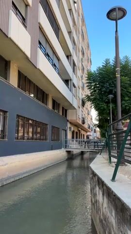 The video shows a view down a narrow canal with water. On the left is a building with multiple stories and windows. On the right is a sidewalk with a fence and a lamppost. In the distance, there is a bridge crossing the canal. The sky is blue and clear. The video is stationary and does not show any movement.