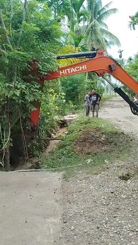 An orange Hitachi excavator is positioned on a dirt road, its boom extended to the left and down, with its bucket appearing to be clearing debris or vegetation from a muddy, eroded ditch. The ditch runs alongside the road, with lush green trees and foliage on the left bank. Two men stand on the right side of the road, observing the excavator's operation. One man wears a light blue shirt and dark pants, while the other wears a dark shirt and pants. Further back on the road, a motorcycle is visible, with another person possibly seated behind the rider. The environment appears to be a rural, tropical setting, with palm trees and dense vegetation visible in the background. The sky is overcast.