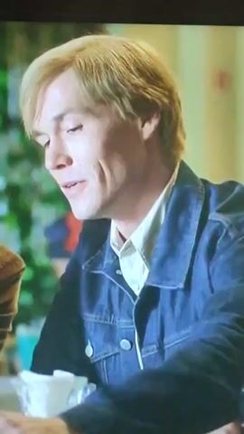 The video displays a close-up of a man with blond hair and a denim jacket smiling and speaking. He is seated at a table with a glass dispenser filled with white napkins. In the background, out of focus, a woman with brown hair and a red scarf is visible, also smiling. The setting appears to be a casual indoor environment, possibly a cafe or restaurant, with green foliage in the background. The man gestures with his hand as he talks. The woman looks directly at the camera.
