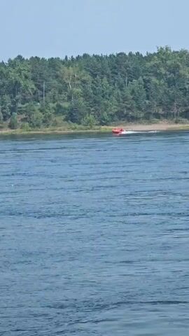 The video shows a wide body of water with gentle ripples. In the distance, a small orange boat with two people on board is moving from left to right across the frame. The boat is creating a white wake behind it. The shoreline is visible in the background, covered with dense green trees. The sky appears to be clear and bright.
