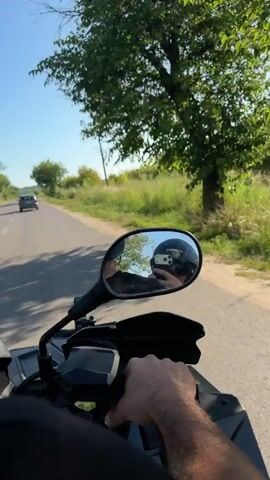 The video is filmed from the perspective of someone riding an ATV on a paved road. The rider's hand is visible on the handlebar. In the rearview mirror, the rider's reflection is visible wearing a black helmet and holding a smartphone to take the video. A dark-colored car is in front of the ATV on the road, and the surrounding environment is rural with trees, bushes, and a power line pole visible against a clear blue sky. The video appears to be captured during daylight.