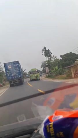 The video shows a view from a vehicle driving on a road with two lanes. There are several trucks and other vehicles on the road. On the left side of the road, there is a blue shipping container truck. On the right side of the road, there is a green fuel truck. There are also some trees and palm trees on the side of the road. The sky is overcast. The camera is positioned inside the vehicle, looking out through the windshield. There are some objects in the foreground that appear to be part of the vehicle's interior, possibly a dashboard and some packaging.