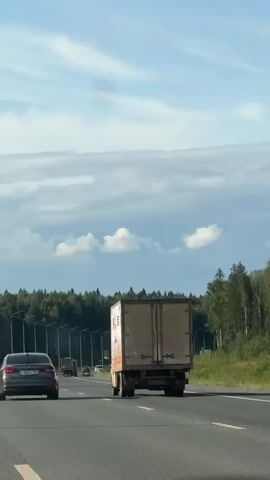 A dark-colored car drives on a highway with a truck in front of it and a forest on the right side of the road. The sky is blue with some clouds.