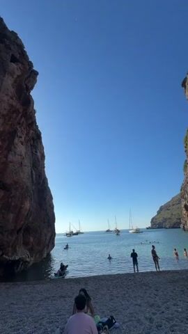 The video shows a pebble beach cove with rocky cliffs rising on either side. Clear blue water stretches out to the horizon, where several sailboats are anchored. People are swimming, wading, and relaxing on the beach. A couple is sitting on a towel, while others stand at the water's edge. The sky is clear and bright.