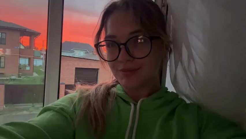 A young woman with round glasses and a green hoodie is sitting inside by a large window. Outside the window, there is a vivid red sunset over a neighborhood of houses with brick and dark-colored roofs. She has her hair in a ponytail. The woman looks directly at the camera with a slight smile. The lighting is dim inside, contrasting with the bright sunset outside. A white curtain is partially visible to the right of the frame.