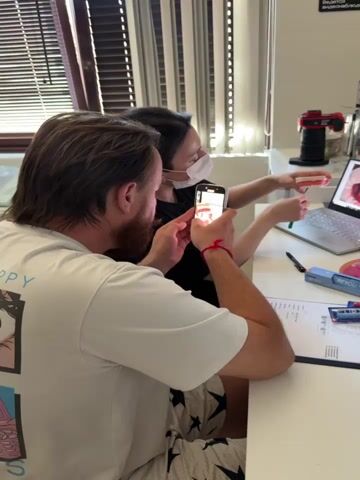 Two people are in a room, likely a dental office or a classroom. A man in a white t-shirt is on the left, holding a smartphone and looking at its screen. A woman wearing a white face mask and a black shirt is on the right, holding a model of teeth and pointing at a laptop screen. The laptop is open and displays an image of teeth. A camera on a stand is visible behind the woman. There are various objects on the table, including pens, a toothbrush box, and papers. The overall environment appears to be a well-lit indoor space with blinds on the window.