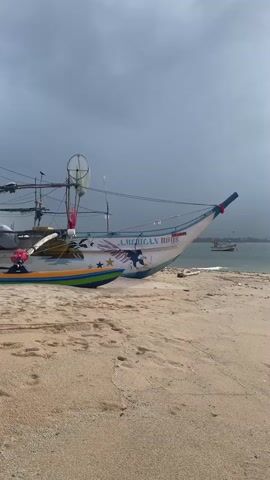 A medium shot captures a traditional fishing boat docked on a sandy beach. The boat, painted white with blue and red accents, displays an eagle graphic and the words AMERICAN BOYS. Footprints are visible on the sand leading away from the boat towards the water. The sea is calm with small waves. The sky is overcast with sunlight breaking through in the upper right quadrant. In the distance, another small boat is visible on the water. The overall atmosphere suggests a tranquil, possibly early morning or late afternoon scene.