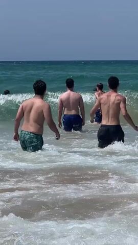 The video shows a group of people in the ocean on a sunny day. The water is choppy with moderate waves. Several individuals, mostly young men, are wading into the water, with some submerged up to their waists. Others are visible further out in the ocean, with some appearing to be swimming or surfing. The scene is set on a beach with a clear blue sky. The focus is on the people and the water.
