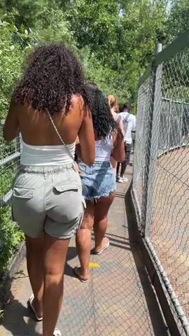 The video shows a group of people walking along a paved pathway. The pathway is bordered by a wire fence on the right and lush green foliage on the left. In the foreground, a hand holds up a cup with a Fanta Frozen drink. The people are walking away from the camera, with the first two individuals being women wearing shorts and tank tops. The environment appears to be an outdoor setting, possibly a park or a zoo, given the presence of greenery and the pathway. The lighting suggests it is daytime and sunny.