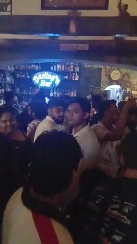 The video shows a crowded bar with a sign that says "Darling's Bar" in bright blue neon lights. There are many people standing and talking.  Behind them is a bar stocked with bottles. The lighting is dim and the atmosphere seems lively.