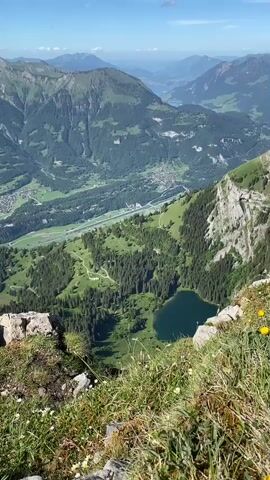The video captures a panoramic view from a mountain peak overlooking a valley with a lake and surrounding mountains. The camera pans across the landscape, revealing lush green slopes dotted with trees and scattered buildings, indicating a rural or mountainous settlement. A winding road or path can be seen traversing the valley. In the foreground, the edge of the mountain is visible, with rocks and grass. At the very end of the video, a hiker's foot wearing a hiking boot is visible in the lower left corner, suggesting the viewpoint is from someone actively hiking or resting at this vantage point. The sky is blue with scattered clouds, indicating pleasant weather. The overall scene is one of natural beauty and expansive mountain scenery.
