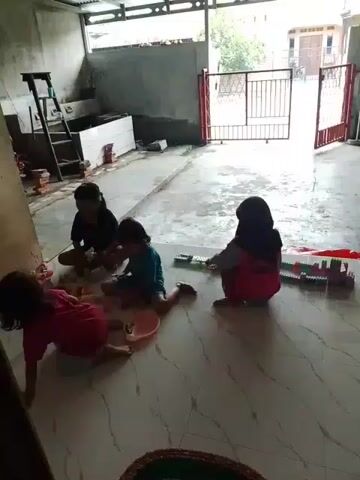 Four children are sitting on a light-colored tiled floor. The child on the far left is wearing a pink shirt. The child next to them is wearing a blue shirt and has their legs extended. The child behind them is wearing a black shirt. The child on the far right is wearing a pink shirt and a black headscarf. They are all playing with colorful building blocks. There is a ladder and some construction materials in the background. The environment appears to be an unfinished or covered outdoor space.