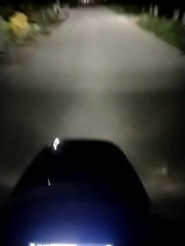 The video shows a first-person perspective of someone riding a scooter at night. The illuminated speedometer displays a speed around 43 km/h. The road ahead is dark but illuminated by the scooter's headlight, revealing a paved surface with streetlights and occasional houses visible in the background. The scooter's black dashboard is also visible in the foreground. There is significant wind noise picked up by the microphone.