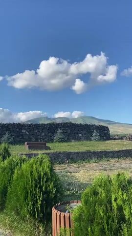 A video captures a wide shot of a rocky mountain range under a bright blue sky with fluffy white clouds. In the foreground, there is a stone wall, a dry, grassy field, and lush green bushes. The video appears to be a panning shot, moving from left to right, showcasing the expansive landscape. The lighting suggests it is daytime.