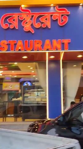 The video captures a street view in Jeddah, Saudi Arabia, from a moving vehicle. The camera pans across the facades of commercial buildings.

Initially, a blue building with brightly lit signs is prominent. One sign features Bengali script, partially visible as "RANT" (likely part of "RESTAURANT"), with the English word "RESTAURANT" also partially visible below it. Another large sign on the same building displays "مطعم" (Mat'am), which means "Restaurant" in Arabic. The restaurant has large glass windows with white curtains inside.

As the vehicle progresses, a second building comes into view, characterized by a green and white sign with Arabic script, possibly reading "مؤسسة عبد الرحمن" (Abdul Rahman Establishment). A person is visible near the entrance of this second building.

Several cars are present on the street: a black car in the foreground, a gray sedan passing by, and a white car briefly seen at the end of the clip. The scene appears to be set during the daytime, under clear weather conditions.