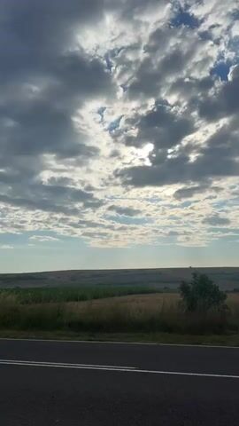The video shows a view of a cloudy sky over a rural landscape. The foreground features a section of an asphalt road with white lane markings. Beyond the road, there is tall, dry grass and a few scattered bushes, with a larger bush slightly to the right of the center. The middle ground reveals a vast agricultural field with rows of crops and patches of dry, golden vegetation, suggesting a harvest season or dry conditions. The horizon is marked by a gentle undulation of the land, with distant fields and trees visible. The sky is filled with a mix of dark, heavy clouds and lighter, scattered clouds, with patches of blue sky peeking through. Sunlight is breaking through the clouds, casting rays of light onto the landscape. The overall impression is one of a rural scene, possibly taken from a moving vehicle on a road, with dramatic cloud formations and a wide-open expanse of fields.