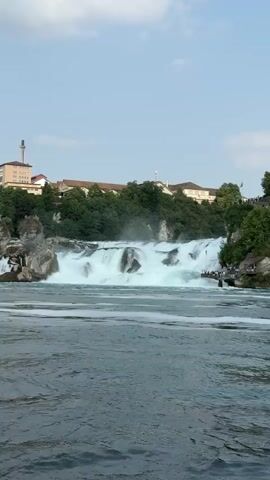 A wide shot captures a powerful waterfall cascading over rocks, creating a misty spray. The water in the foreground is choppy with whitecaps. A tour boat is visible on the left side of the frame, moving across the water. On the right side of the waterfall, a wooden walkway is lined with small groups of people observing the falls. Behind the waterfall, lush green trees and buildings are perched on a hill under a clear blue sky with a few wispy clouds. The scene appears to be a scenic tourist destination on a pleasant day.