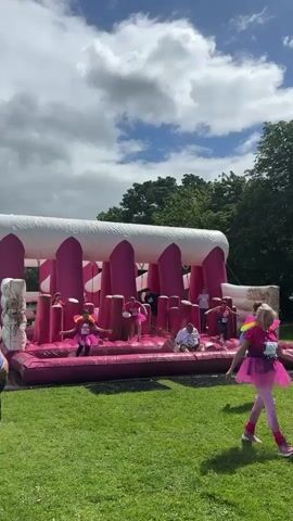 The video shows an outdoor scene with an inflatable obstacle course in the center. The obstacle course is predominantly pink and white and appears to be set up on a grassy area. Several people are interacting with the obstacle course, some are climbing or navigating through it, while others are standing or walking around it. One person is on the ground within the obstacle course, appearing to be crawling or getting up. Another person in a blue shirt and shorts is standing outside the obstacle course facing it. The sky is cloudy with patches of blue visible. Trees are present in the background. The people are wearing casual clothing, and some are wearing pink tutus. The overall impression is of a fun, active event.