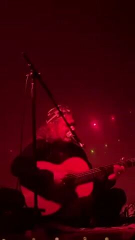 The video shows a musician sitting and playing a classical guitar on stage under red lighting. A microphone stand sits to his left. The musician has long, curly hair and wears a patterned headband. He is wearing a black shirt. There are green and red laser dots in the background. A dark figure briefly crosses the bottom left corner of the screen at the end.