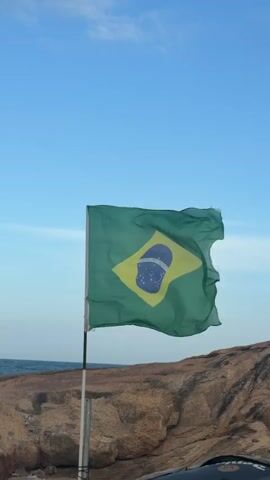 A Brazilian flag flutters in the wind against a clear blue sky. The flag is green with a yellow rhombus in the center, containing a blue circle with stars and a curved white band. The flag is attached to a metal pole, which is partially visible. In the background, a rocky coastline meets the ocean, with waves gently lapping at the shore. The lighting suggests it is daytime, likely late afternoon or early morning due to the soft light. There are no people visible in the frame.