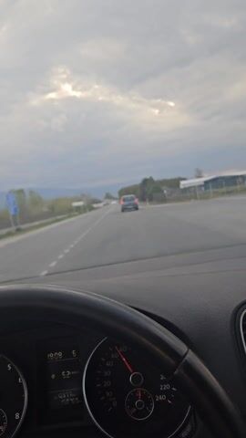 The video shows a car driving down a highway on a cloudy day. Another car is in front, traveling in the same direction. The dashboard is visible with a speedometer reading approximately 52 km/h. The sky is overcast, but some light is shining through the clouds. Trees and buildings can be seen on the side of the road.