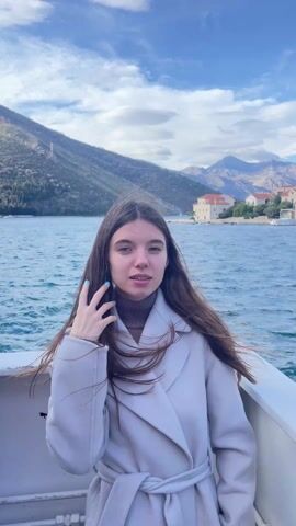 A young woman with long brown hair stands on a boat, facing the camera. She is wearing a light gray coat and a brown turtleneck sweater. The wind is blowing her hair. Behind her is a body of water, with mountains in the background. There are some houses visible on the shore. The sky is blue with some clouds. The woman is looking off to the side and touching her hair with her hand.
