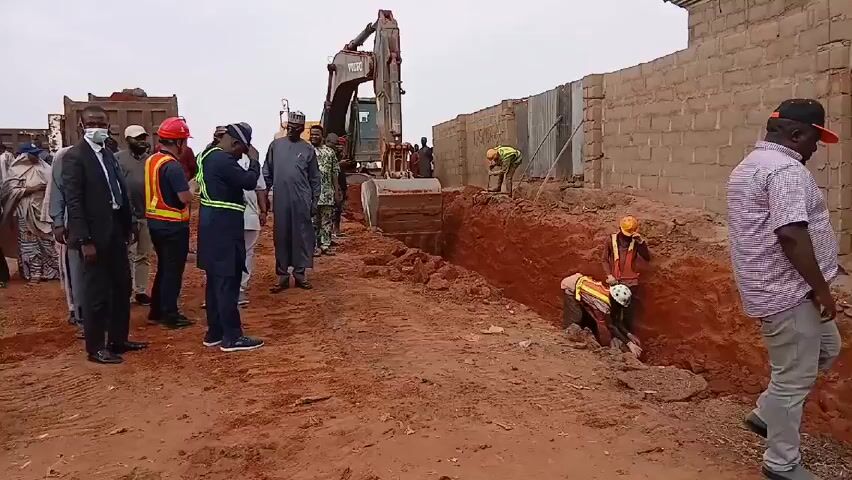 A group of people are standing near a trench being dug by a large excavator. Some of the people are wearing business attire, while others are wearing hard hats and safety vests. There is a brick wall on one side of the trench and a dump truck in the background. The ground is reddish-brown. Several workers are inside the trench, seemingly working on something.