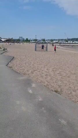 The video captures a beach scene on a sunny day, with a blue sky and scattered clouds overhead. A grey concrete pathway is visible on the left side of the frame. The beach itself is sandy, with footprints and marks visible in the sand. Three children are playing in the center of the frame near a cage-like structure constructed from metal bars. One child is in black clothing, while another wears a red shirt. The third child is wearing green. The child in the red shirt falls to the ground. In the background, people can be seen along the beach, and there is a view of a city skyline in the distance, along with boats docked in a marina. The scene is relatively static, with the primary action focused on the children playing near the cage structure.