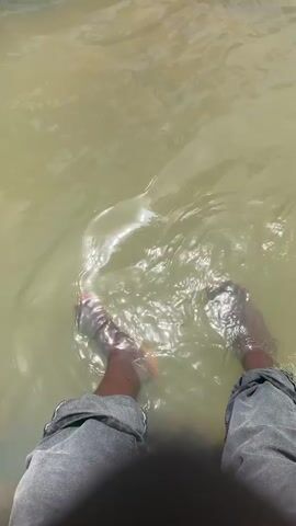 This media file captures a close-up perspective of a person's legs and feet submerged in murky, greenish-brown water. The water appears shallow and is rippled, suggesting movement or disturbance. Sunlight glints off the surface, creating bright highlights. The person is wearing grey denim shorts with visible stitching and a dark, out-of-focus element obscures the lower torso. The individual's skin tone is dark. The feet are visible, with toes slightly splayed. The scene appears to be outdoors, given the natural light. The activity is simply the person having their feet in the water. There is no visible text in the media file. The setting might be a natural body of water such as a river, pond, or possibly a shallow, unmaintained pool, given the water's appearance. The mood conveyed is relaxed and possibly summery or warm weather.