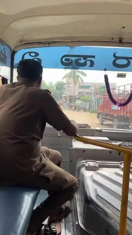 A medium shot shows the interior of a bus from the perspective of a passenger. A male bus driver, wearing a brown shirt and pants, is seated in the driver's seat, facing forward. He is holding the steering wheel with his left hand, and his right hand is near the gear shifter. The bus is stationary or moving slowly, as indicated by the visible exterior scene. Through the windshield, a dirt road is visible, with buildings and trees on either side. A red tractor with a trailer is parked on the side of the road. There is a red truck to the right, and a green structure is visible behind it. The windshield is decorated with a purple garland and some text in an unknown script. Another person, possibly a passenger, is partially visible in the foreground, leaning on a yellow handrail. The interior of the bus appears functional and somewhat worn. The lighting suggests it is daytime.