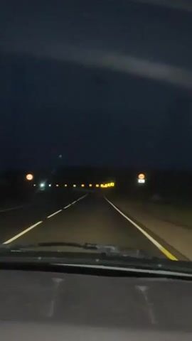 This is a first-person perspective video of driving on a highway at night. The road has two lanes in each direction, separated by a solid white line. The sky is dark and appears cloudy. There are numerous yellow, illuminated signs on the right side of the road, some of which indicate curves. On the left side, there are also illuminated signs. The headlights of the vehicle are on, illuminating the road ahead.