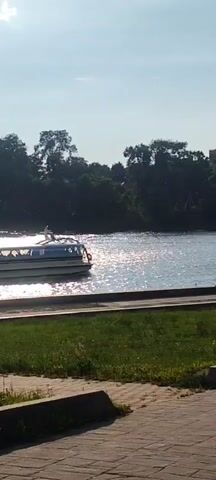 A white and blue passenger boat travels along a wide body of water. The boat has a white upper deck with windows. Several people are visible on the rear open-air deck of the boat. The water reflects the bright sunlight, creating a shimmering effect. On the left side of the frame, a line of trees forms a dark backdrop against a light blue sky. In the foreground, a strip of green grass is bordered by a paved walkway.
