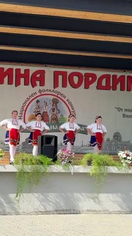 The video shows a group of five young girls performing on an outdoor stage. They are dressed in traditional folk costumes, consisting of white blouses with red and blue accents, and blue skirts with red aprons. Each girl has her hair styled in braids. They appear to be dancing or performing as part of a cultural event.

The stage has a backdrop with large red lettering that reads "ПОРАДИМ" (PORDIM). There are also circular logos and text in Cyrillic script on the backdrop, suggesting a folk competition or festival. One of the visible texts translates to "From the cradle of the North" and "Municipality Pordim". Another quote reads "From a small place - a great deed."

The stage is decorated with green foliage and floral arrangements in white and pink. There are speakers visible on the stage. The lighting suggests it is daytime, likely sunny, given the bright illumination and shadows. The overall atmosphere appears festive and celebratory, focusing on traditional Bulgarian culture. The girls' expressions are difficult to discern clearly, but their posture and movements suggest engagement in their performance.