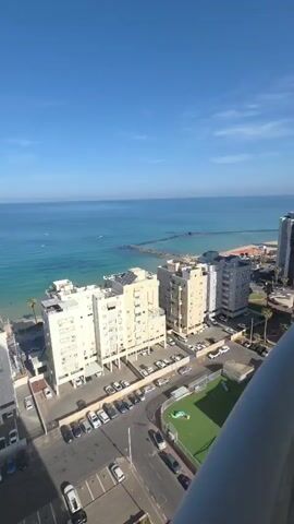 The video presents a high-angle view of a coastal scene, likely taken from a tall building. The focus is on the expanse of turquoise-blue ocean under a clear, bright blue sky with minimal clouds. In the middle distance, there are a few small boats on the water.  Closer to the shore are several white buildings, a parking lot filled with cars, and a small green sports field.  There is a pier extending into the ocean. A shadow from the building the video is being taken from is also visible on the left side of the scene. The video zooms in on a couple of yellow kayaks on the water.