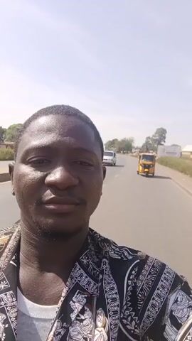 A man is taking a selfie-style video while on a motorcycle. He is wearing a patterned shirt and faces the camera with a neutral expression. The road stretches behind him with sparse traffic consisting of a white van, a car, and a yellow auto-rickshaw in the distance. The weather appears sunny, and the sky is clear. Trees and buildings can be seen alongside the road. At the 0:00 mark, an explosion sounds briefly.