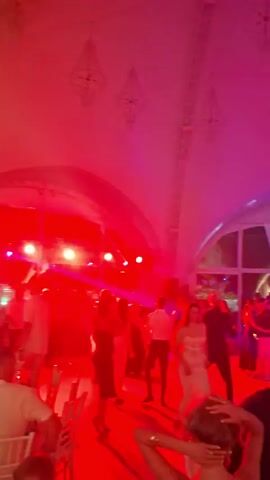 The video shows a group of people dancing indoors under colored lights that shift from blue to red to green and back to blue. The room appears to be a banquet hall or event space with a high, arched ceiling and large windows in the background. Several people are visible on a dance floor, with some seated at tables in the foreground. The lighting creates a festive atmosphere, with beams of light cutting through the air. The overall impression is of a lively party or celebration.
