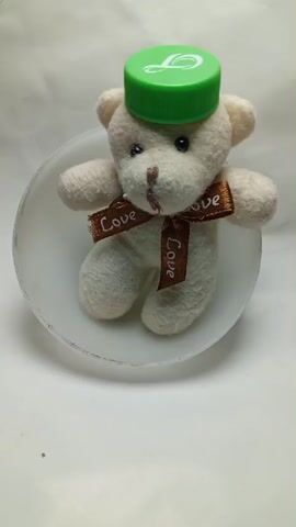 A small, cream-colored teddy bear is positioned inside a translucent bowl. The bear is wearing a green bottle cap as a hat, and a brown ribbon with the word "Love" printed on it is tied around its neck. The background is a plain white surface. The video shows a static shot of the teddy bear in the bowl.