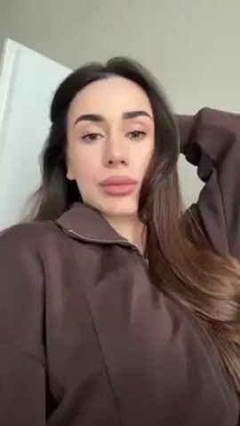 A woman lies on a light-colored couch. She is wearing a brown, long-sleeved, collared sweatshirt with a zipper. Her long brown hair is spread out on the couch. The wall is white. The woman smiles.