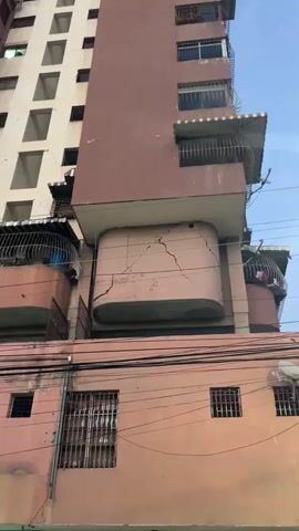 The video shows a close-up view of a tall, aging apartment building. The building appears to be in a state of disrepair, with visible cracks on its exterior walls. The walls are painted in muted colors, primarily a pale beige and a dusty rose. The windows are mostly barred, suggesting a need for security, and some appear to be empty openings.

In the foreground, electrical wires crisscross horizontally, indicating an urban setting. Balconies with metal railings are visible, and one balcony has a large, curved metal cage or enclosure on it, with a blue object inside. The overall impression is one of a densely populated urban environment where maintenance may be a challenge.

The camera pans slowly upwards, revealing more of the building's facade and the extent of the cracks. The time of day is difficult to ascertain precisely, but the lighting suggests it might be dusk or a cloudy day, as there are no strong shadows. There are no people clearly visible in the footage, though the presence of balconies and windows implies inhabitants.

The scene is static, focusing on the structural condition of the building. The cracked walls and general wear and tear evoke a sense of neglect and potentially concern for the building's stability. The location context of Maracay, Venezuela, provides a geographical anchor to this visual documentation of urban infrastructure. No legible text is visible in the video.
