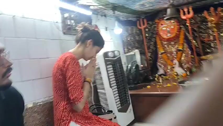 The video depicts a scene inside what appears to be a small Hindu temple. A woman in a red and white patterned kurta is kneeling and praying with her hands together. A man in a white undershirt and red pants sits near her, facing the altar with a deity image. The altar is adorned with golden accents, including tridents, swan statues, and hanging decorations. Another man, wearing a black jacket and glasses, is close to the seated man, who is applying a red mark on his forehead. There is a white tower fan and a small oscillating fan in the background to the left of the altar. The environment is enclosed, with white tiled walls and a wooden cabinet forming the base of the altar. The scene suggests a religious ceremony or a visit for blessings at the temple.